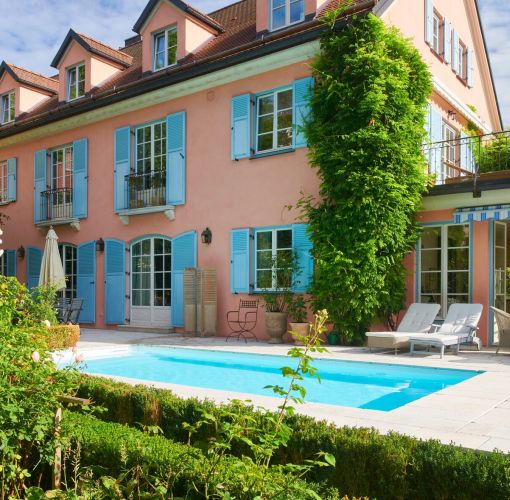 Haus mit blauen Fensterläden und Pool im sonnigen Garten, Hecke und Kletterpflanzen im Vordergrund